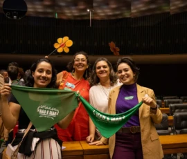 Foto com integrantes da campanha Criança Não é Mãe. Duas mulheres seguram lenços verdes. Atrás delas, outras duas seguram flores de papel em amarelo e laranja, símbolo da campanha Faça Bonito.