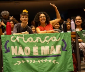 Foto da bandeira da campanha Criança Não é Mãe e sentada atrás de uma bancada, estão duas ativistas, uma delas tem o braço levantado. Atras delas outras pessoas estão com a mão levantada.