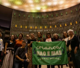 Foto de integrantes da campanha Criança Não é Mãe segurando a bandeira verde da campanha. Atrás, um painel e luzes do plenário da Câmera Federal.
