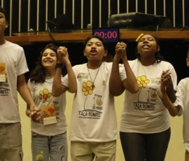 Foto com cinco crianças de mãos dadas erguidas, cantando. Elas estão com a blusa da campanha Faça Bonito.