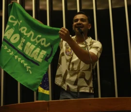 Foto de um parlamentar discursando no pulpito do plenário da Câmera Federal. Ele ergue a bandeira da campanha Criança Não é Mãe.