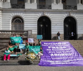 Natália Elmor Foto de ativistas sentadas nas escadarias da Câmera de Vereadores do Rio de Janeiro, erguendo lenços, cartazes e bandeiras da campanha Criança Não é Mãe. À frente, uma coroa de flores e à direita uma bandeira grande roxa com a frase "Ninguém deve ser maltratada, presa ou humilhada por ter feito aborto! Frente Estadual Contra Crimanilização das Mulheres e pela Legalização do Aborto".