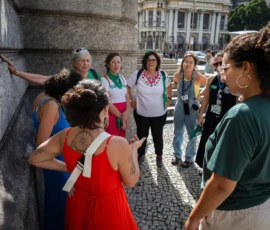 Natália Elmor Foto de um grupo de mulheres em diálogo, reunidas em círculo. Atrás delas um prédio histórico.