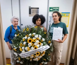 Natália Elmor Foto das ativistas Mariane Marçal, Laura Molinari e Angela Freitas, na qual uma segura a coroa de flores com a frase "Em luto frente à Casa que lesgila pela morte de mulheres, meninas e pessoas que gestam", outra mostra uma carta. Atrás delas, placas e cartazes na parede, um deles diz: Gabinete Vereador Carlo Caiado.