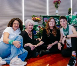 Foto com as quatro integrantes da equipe Nem Presa Nem Morta, sentadas ao chão. Atrás delas, o cenário Altar Feminista e percianas brancas fechadas.