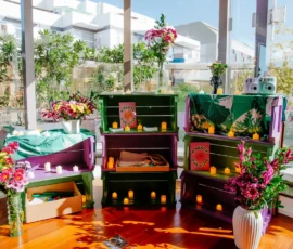 Foto de um cenário, chamado de Altar Feminista, com caixas verdes e roxas empilhadas. Nelas estão distribuídas velas, decorações e plantas. Atrás, uma parede de vidro com plantas no espaço externo.