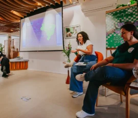 Foto das palestrantes Lucilene e Laura Molinari sentadas à frente do flip chart. Ao lado, uma mesa pequena com um copo de água e uma planta. Em seguida, um telão onde está reproduzida a imagem de um mapa do Brasil, dados e informações. Sentada em frente está Angela Freitas, mediadora da atividade.