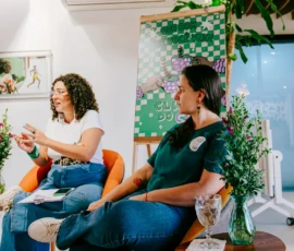 Foto das palestrantes Lucilene e Laura Molinari sentadas. Ao lado de cada uma tem uma mesa pequena com um copo de água e uma planta. Atrás, um flip chart e um quadro.