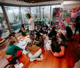Foto das participantes sentadas no chão fazendo atividades de cortar, colar e desenhar sobre cartazes. Espalhados no piso da sala estão canetas, linhas, revistas e outros materiais. Ao centro da foto, em pé, uma das partipantes fotografa um cartaz. Na lateral também há pessoas em pé. Ao fundo um painel com cartazes coloridos da Nem Presa Nem Morta e portas grandes de vidro.