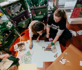 Foto com ângulo alto de duas participantes sentadas ao chão, debruçadas sobre um cartaz no qual colam recortes de revista. Atrás delas, aparece parte do cenário com caixotes roxos e verdes, lenços, flores e velas do Altar Feminista.