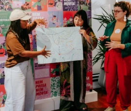 Foto de três participantes aprensentando um cartaz que contém linhas coloridas e desenhos de mãos. Do lado esquerdo, uma delas está de bonè e gesticula sobre a imagem enquanto a outra segura o cartaz. No canto direito, a terceira participante também gesticula com as mãos. Atrás delas, um painel colorido com cartazes da Nem Presa Nem Morta.
