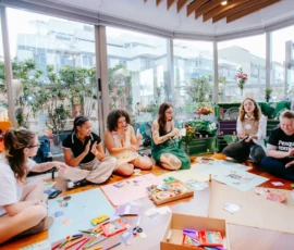 Foto das participantes sentadas fazendo atividades de cortar, colar e desenhar. No chão estão cartazes coloridos, lápis, linhas, tesouras, caixas e outros materiais. Atrás delas portas grandes de vidro.
