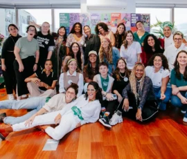Foto das participantes, convidadas e equipe posando e sorrindo para câmera. A maioria está em pé, algumas agachadas e duas delas deitadas. Atrás, um painel colorido com cartazes da Nem Presa Nem Morta e portas grandes de vidro.