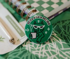 Foto em close de um boton verde redondo com a ilustração de uma gata, acima a frase "Deixa a gata decidir". Ao lado, aparece parte de um lápis e de um caderno com aspiral dourado.