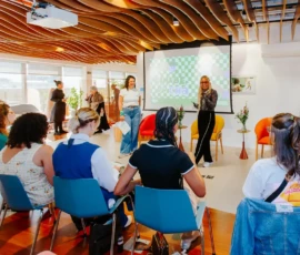 Foto da sala em que seis participantes estão sentadas, de costas para a câmera, em cadeiras azuis. Ao fundo da imagem estão em pé as apresentadoras da atividade. Atrás delas, poltronas, objetos decorativos e um telão com a logo Clube do Chá.