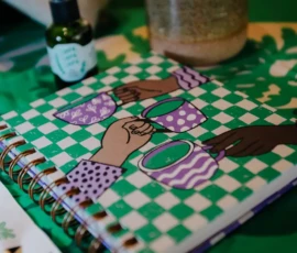 Foto de um caderno com aspiral dourado. Na capa, em quadriculado verde e branco, tem a ilustrações de três mãos segurando xícaras de chás com estamparia roxas e brancas. Em volta do carderno elementos desfocados.