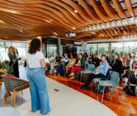 Foto ampla da sala com primeiro plano nas apresentadoras em pé, de costas para a câmera. À frente delas, participantes assistem as falas sentadas. Teto com estacas de madeira caramelo em formato ondulado.
