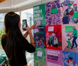 Foto com uma jovem de costas para câmera. Ela registra no celular a imagem de um postit que está colado em um painel grande com cartazes coloridos da Nem Presa Nem Morta.