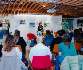 Foto ampla da sala em que as participantes estão sentadas de costas para câmera. Ao fundo, Bibiane Serpa está em pé e segura um caderno. Logo atrás, poltornas e quatro quadros na parede. Ao lado, um flip chart com a logo do Clube do Chá.