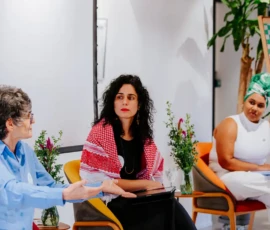 Fotos das três palestrantes sentadas, da direita para esquerda: Mariane Marçal, Mariana Prandini e a Pastora Lusmarina Campos, que gesticula com as mãos. Ao lado delas, aparece parte do flip chart com a logo do Clube do Chá.