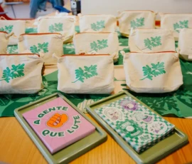 Foto de uma mesa com várias nécessaires em tecido de algodão cru com a gravuras de ramo de arruda verde. À frente, dois blocos empilhados com: adesivos e outro de papel cartão, com a frase "A gente que lute". Uma canga estampada em verde da Nem Presa Nem Morta forra a mesa.