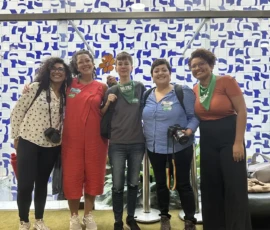 Foto de cinco comunicadoras com lenços e adesivos da campanha Criança Não é Mãe no roll do Câmara Federal. Duas delas seguram câmeras fotográficas. Atrás delas, uma parede de azuleijos estampados em brancos e azuis.