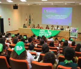 Foto ampla de auditório cheio, com visão das costas do público. Ao fundo, a bandeira grande da Nem Presa Nem Morta e uma mesa com dez palestrantes. Atrás delas, um telão com a logo e nome do encontro.