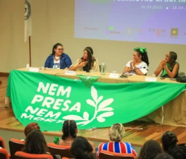 Foto da mesa com quatro palestrantes sentadas atrás da mesa com uma grande bandeira da Nem Presa Nem Morta estendida.