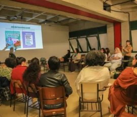 Foto de sala com participantes sentadas em meio cículo, assistindo a apresentação projetada na parede do conteúdo da oficina "Comunicação para pesquisadoras sobre aborto" da Nem Presa Nem Morta.