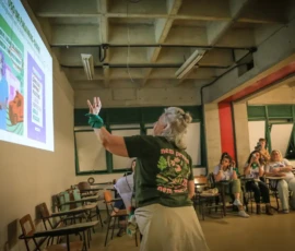 Foto de Angela Freitas de costas, apresentando o conteúdo da oficina "Comunicação para pesquisadoras sobre aborto" da Nem Presa Nem Morta. À sua esquerda, uma projeção na parede.