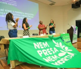 Foto de quatro palestrantes aplaudindo, em pé, no auditório. Na frente delas, uma mesa com uma grande bandeira da campanha Nem Presa Nem Morta e outra menor da campanha Criança Não É Mãe.
