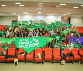 Foto ampla do auditório, onde diversas pessoas estão em pé erguendo lenços verdes e bandeiras.
