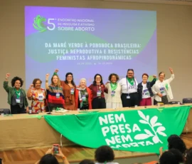 Foto das dez palestrantes em pé, abraçadas. Elas estão por trás de uma mesa na qual está estendida uma grande bandeira da Nem Presa Nem Morta. Atrás delas, um telão com a logo e nome do encontro projetada.