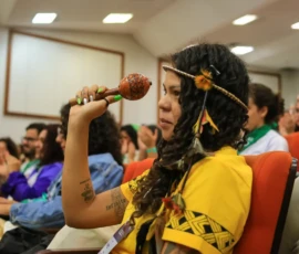 Foto em close de uma mulher indígenas com penacho nos cabelos e maracá na mão.