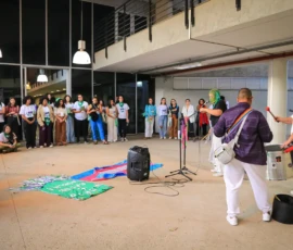 Foto do público assistindo a uma intervenção artística de ativistas com tambores e microfones. No chão, a bandeira trans e um estandarte verde estendidos.