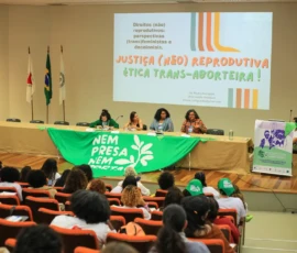 Foto ampla com parte do público de costas e a mesa com quatro palestrates. Na frente da mesa há uma grande bandeira da Nem Presa Nem Morta. Atrás, um telão com uma apresentação projetada: Justiça (não) reprodutiva, ética trans-aborteira!