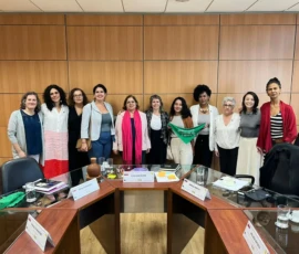Acervo Ministério das Mulheres Foto da comitiva de mulheres com a Ministra Cida Gonçalves. Na frente delas, cadeiras e a mesa da reunião. Atrás, uma parede em madeira.