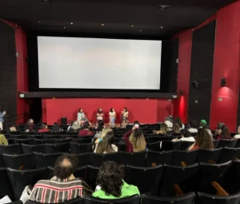 Foto de uma sala de cinema com as luzes acessas, tela apagada, plateia nas poltronas e quatro palestrantes sentadas de frente para o público.