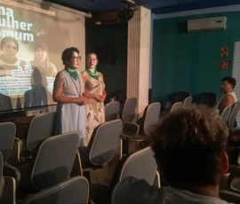 Foto de duas mulheres com lenços verdes no pescoço, apresentando o filme "uma mulher comum" para uma pequena plateia em um auditório. Atrás delas, a imagem do cartaz do filme.