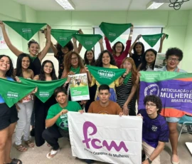 Foto de pessoas posando com banideras e lenços verdes da campanha Criança Não é Mãe em uma sala.