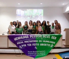 Em um grande auditório, pessoas posam pra foto atrás de uma bandeira grande com o texto: Nenhuma pessoa deve ser presa, maltratada ou humilhada por ter feito aborto. Frente Nacional Contra a Criminalização das Mulheres e pela Legalização do Aborto. Ao fundo, o telão com imagem do filme "uma mulher comum".
