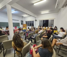 Foto de pessoas de costas sentadas em cadeiras em uma sala. Ao fundo, uma mesa com palestrantes.