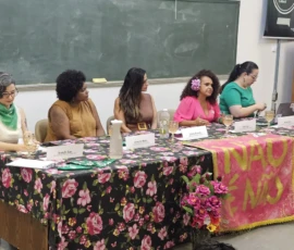 Foto de cinco participantes sentadas em uma mesa forrada com tecido de flores e ao centro uma bandeira escrita "não é não".