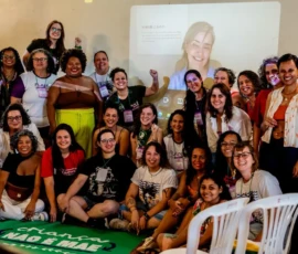 Mulheres posando para foto, algumas em pé outras sentadas no chão. Na frente delas, a bandeira da campanha Criança Não É Mãe. Atrás, a projeção da vídeo-chamada com a diretora do filme Débora Diniz.