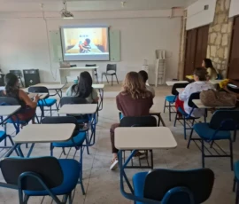 Foto de pessoas sentadas em carteiras universitárias, em uma sala. Ao fundo, uma tela com o filme "uma mulher comum"