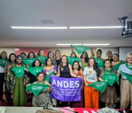 Mulheres posando para foto com lenços verdes e uma bandeira escrita "Andes - Sindicato Nacional", em um auditório.